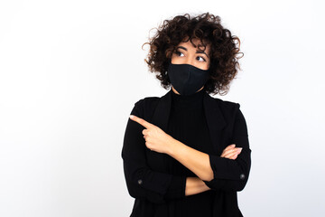 Portrait of young beautiful caucasian woman wearing medical mask standing against white wall, posing on camera with tricky look, presenting product with index finger. Advertisement concept.