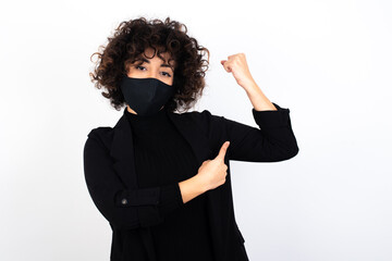 Smiling young beautiful caucasian woman wearing medical mask standing against white wall, raises hand to show muscles, feels confident in victory, strong and independent.