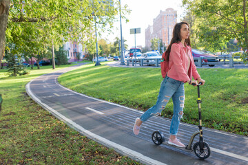 Woman on a scooter, in summer in city rides in motion, free space for a copy of text. Pink jacket, backpack, jeans. Background green lawn, trees, bike path, road and cars.