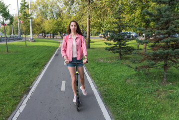woman in summer in city rides a scooter on a bicycle path, in motion, background road trees green grass and spruce, pink jacket denim shorts. Free space for a copy of the text.