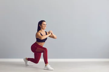 Fototapeten Smiling fitness trainer showing how to do fitness exercise. Happy fit young woman in sports bra and leggings doing forward lunges holding hands together in front of chest during workout at the gym © Studio Romantic
