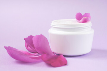 Container with white moisturizing cream for skin care with peony extract and petals on pink background