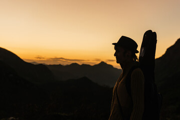 silhouette of female guitarist with hat walking up the mountain carrying her guitar at sunset. musical concept