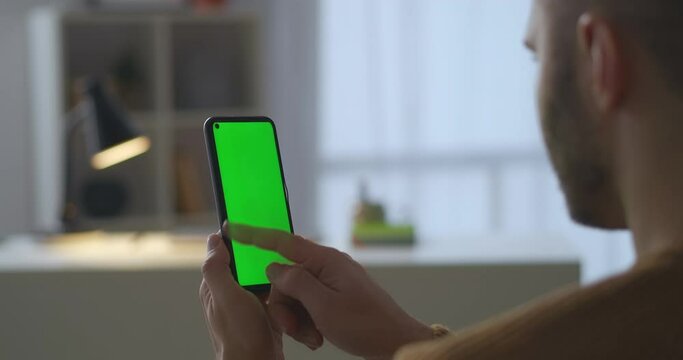 male user is swiping display of smartphone with green screen for chroma key technology, viewing pictures and photos in social nets