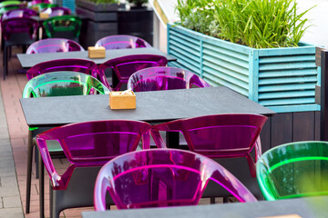 a wooden table top with a napkin holder and plastic multi-colored chairs on the terrace of a street cafe with free seats, nobody.