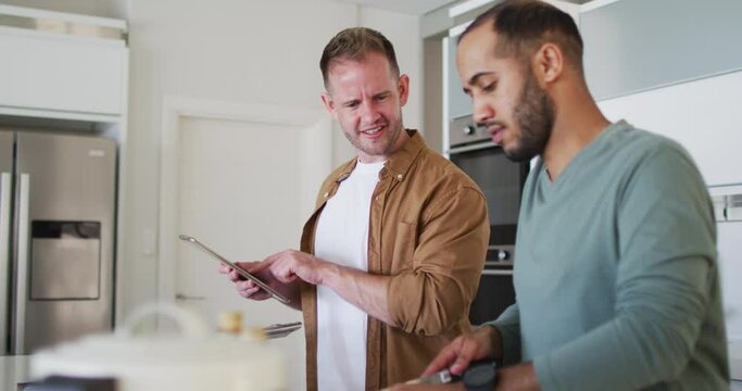 Multi ethnic gay male couple preparing food in kitchen one using tablet