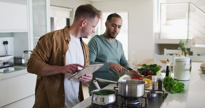 Multi ethnic gay male couple preparing food in kitchen one using tablet