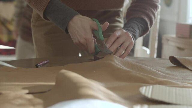 Locked down shot of a person carefully running big scissors through brown thick leather placed on table for precision, to make leather moccasins