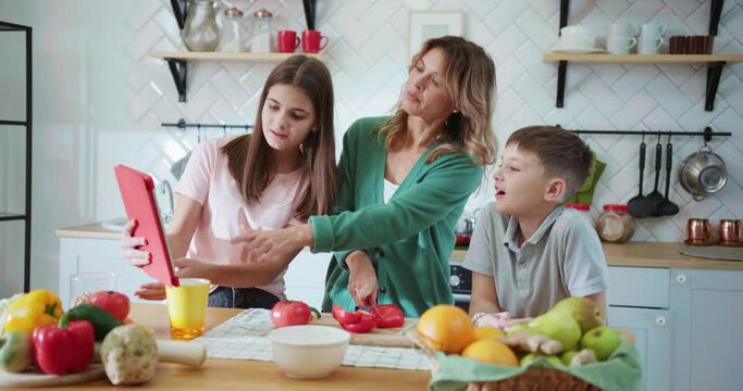 Affectionate family cooking breakfast in the kitchen. Happy blonde mom slicing vegetables for fresh breakfast salad and talking to her children.