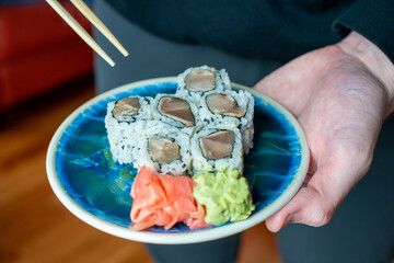 A small side plate of sushi on a decorative blue plate.  The plate has ginger and wasabi on the side. The rolls have tuna, salmon and crab along with seaweed and rice. They have pieces of avocado too.