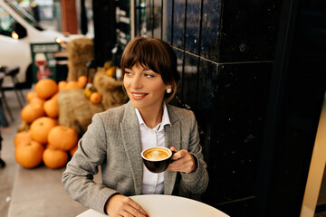  Pretty lady in grey jacket and white shirt widely smiling and drinking coffee. Cool woman is...