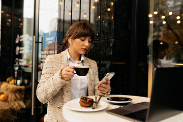 Seriously young stylish business woman in jacket using smartphone and working on laptop, listening music and having coffee break 