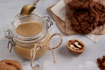 Vegetarian traditional breakfast close up. Glass jar with natural walnut paste, golden spoon, healthy cookies, walnuts on a light background