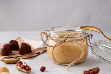 Glass jar with natural peanut butter, golden spoon, peanuts and sweets on a light background. Delicious breakfast close-up. Copy space