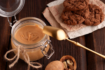 Glass jar with natural nut butter, golden spoon, healthy cookies, walnuts on a brown wooden background. Vegetarian traditional breakfast close up.