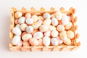 Mixed chocolate coated Easter eggs candies in a small squared wooden basket isolated on a white table, tasty sugary dessert for children.