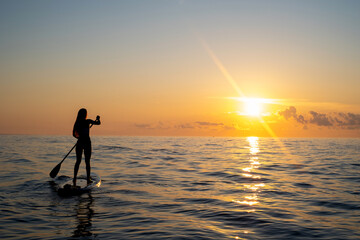 a girl with a paddle on a sub board floats towards dawn on the water in the ocean. Healthy lifestyle concept, water sports. Beautiful sunrises