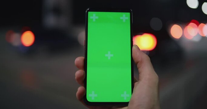 POV, male hand holding smartphone mockup green screen at the night city on bridge, car traffic background
