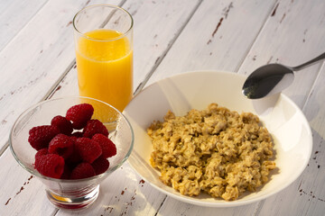 Breakfast: oatmeal, raspberries and orange juice on a white wooden table