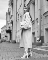 Elegant woman in white dress, hessian boots and coat walking at city street. Fall autumn fashion look. Pretty tall stylish young girl with fashionable makeup and hair style. Full length portrait