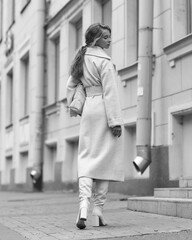 Elegant woman in white dress, hessian boots and coat walking at city street. Fall autumn fashion look. Pretty tall stylish young girl with fashionable makeup and hair style. Full length portrait