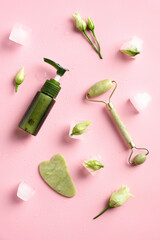 Jade stone face roller, gua sha, facial lotion and ice cubes with flowers on pink background. Top view, flat lay. Facial skin care concept.