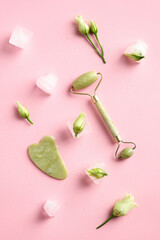 Gua Sha massage tool, jade stone face roller on pink background with ice cubes and flowers. Top view, flat lay.