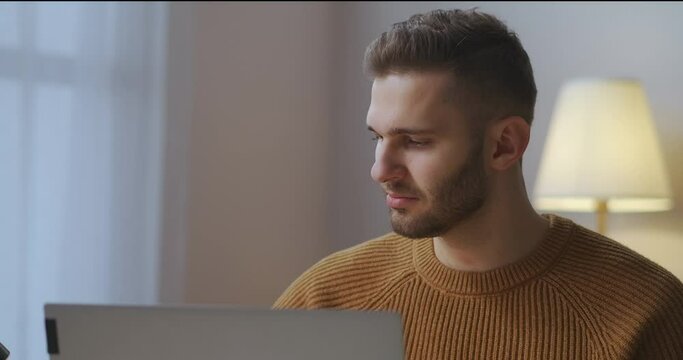male user is working with laptop and putting on glasses for correction vision, surfing internet at evening at home