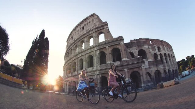 Three happy young women friends tourists riding bikes at Colosseum in Rome, Italy at sunrise.