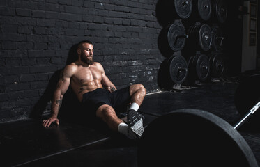 Young active strong fit sweaty muscular man sitting on the floor of the gym after barbell weight...