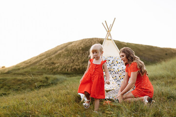 Beautiful mother with her little daughter sitting near wigwam in the field. Spending time together, outside, on vacation, outdoors. Beautiful sunset light in the garden or in the park © Andriy Medvediuk