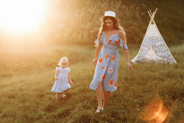 Beautiful mother with her little daughter near wigwam in the field. Spending time together, outside, on vacation, outdoors. Beautiful sunset light in the garden or in the park. © Andriy Medvediuk