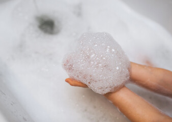 Girl, child holding soap foam in hands over a bath with water.