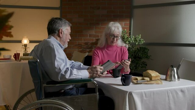 A disabled man in a wheelchair sits in a restaurant with his lady friend talking and laughing as they share images on their digital devices.