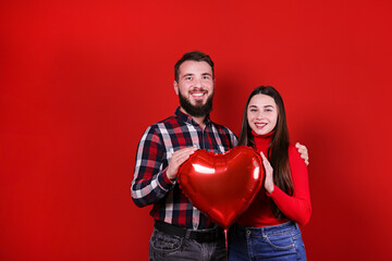 Happy Valentine's day concept. Studio shot of couple wearing casual outfits with heart shaped balloon. Portrait of hipster bearded guy and girl showing affection. Red wall background, copy space