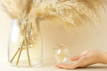 Pampas grass in glass vase, human hand holding transparent glass egg on beige background. Easter holiday, modern minimal composition. Eco Stylish decor concept.