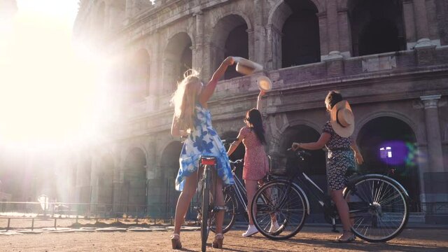 Three happy young women friends tourists with bikes waving hats at Colosseum in Rome, Italy at sunrise.