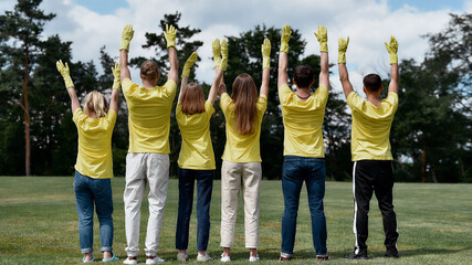 Charity work. Group of young people, volunteers wearing uniform and rubber gloves raising hands up,...