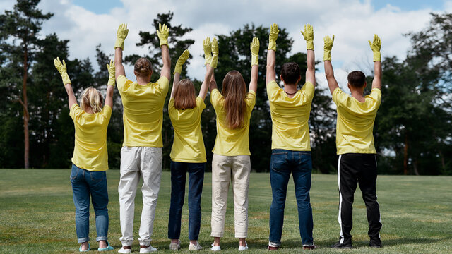Charity work. Group of young people, volunteers wearing uniform and rubber gloves raising hands up, celebrating success after cleaning forest or park together