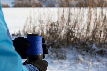 A man holds a thermo mug with hot beverage against the backdrop of a winter river. Concept of winter outdoor recreation. Copy space. Horizontal orientation.