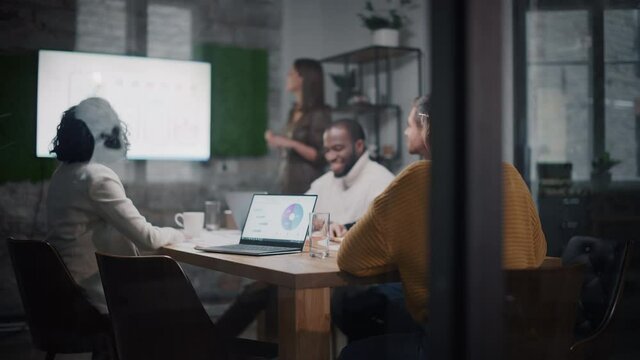 Project Manager Makes a Presentation for a Young Diverse Creative Team in Meeting Room in an Agency. Colleagues Sit Behind Conference Table and Discuss Business Development, Sales Analysis and Design.