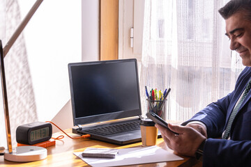businessman at his desk with laptop and coffee working from home and smiling at what is sent to him on his mobile phone