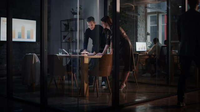 Two Young and Successful Colleagues Have a Conversation in a Meeting Room Behind Glass Walls in an Agency. Beautiful Female Director and Handsome Project Manager Discuss Work on Laptop Computer.