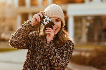 Blissful female model in elegant coat posing with camera. Smiling photographer taking pictures of autumn city.