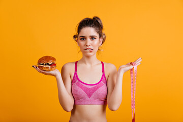 Hesitating woman holding cheeseburger. Studio shot of caucasian girl on diet.