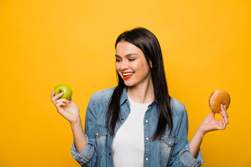 Fast food or healthy food. Caucasian attractive brunette girl holds an apple in one hand and in the other hamburger, making a choice, standing on isolated orange background