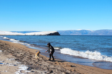 Sunny frosty day. A young woman in warm clothes plays on the shore of Lake Baikal with a dog. The sandy beach is covered with snow. Snow-capped mountains and hills in the background.