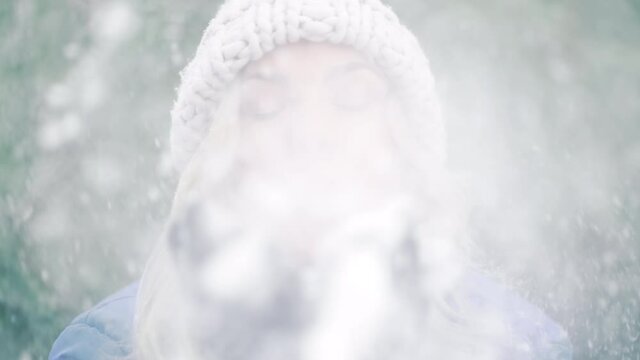 Happy woman blowing snow from palms with mittens to camera on winter forest background. Portrait of cute girl childishly rejoices first snow.