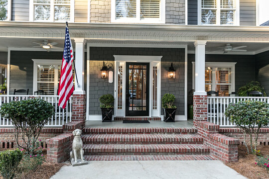 Front Door Entrance To A Large Two Story Blue Gray House With Wood And Vinyl Siding And A Large American Flag.