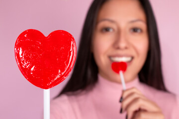 Young woman smiles and eats red heart shaped candy and shows another red heart shaped candy, black hair, pink t-shirt and pink background. She is out of focus and the candy in focus.